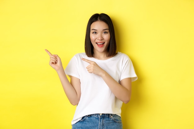 Conceito de beleza e moda. Linda mulher asiática em t-shirt branca apontando os dedos para a esquerda, em pé sobre um fundo amarelo.