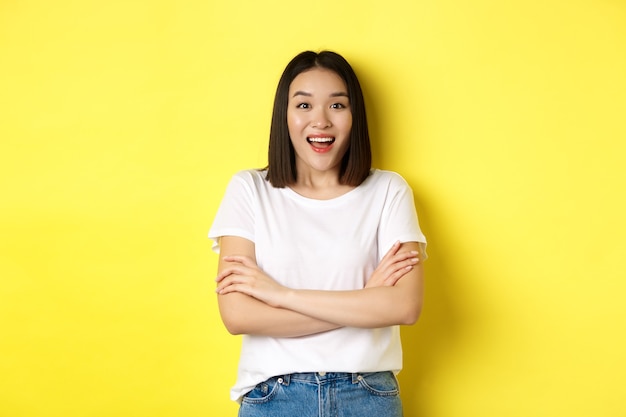 Conceito de beleza e moda. Mulher asiática feliz em uma camiseta branca, braços cruzados no peito, olhando maravilhada com a câmera, em pé sobre um fundo amarelo