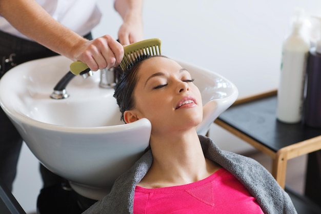 Conceito de beleza e pessoas - jovem feliz com cabeleireiro penteando o cabelo molhado após lavar no salão