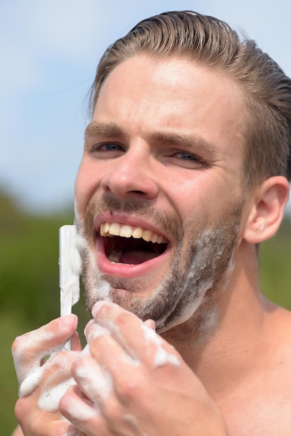 Conceito de beleza e saúde. Homem com cerdas no fundo do céu claro. Macho raspando o queixo com navalha. Cara coberto com espuma de barbear. Macho com rosto sexy.