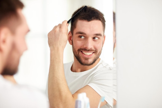 Foto conceito de beleza, higiene e pessoas - jovem sorridente olhando para o espelho e escovando o cabelo com pente no banheiro de casa