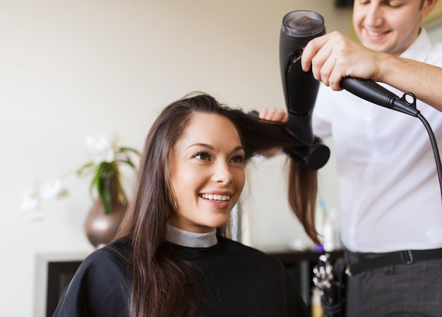 conceito de beleza, penteado e pessoas - jovem feliz e cabeleireiro com ventilador fazendo estilo quente no salão de cabeleireiro