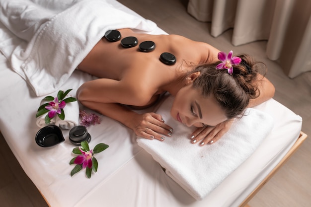 Conceito de bem-estar, beleza e relaxamento - mulher jovem e bonita com massagem com pedras quentes no spa