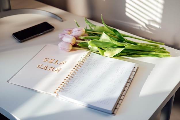 Conceito de bem-estar de autocuidado com flores de tulipa de palavra de autocuidado de notebook aberto na mesa branca, cuide-se