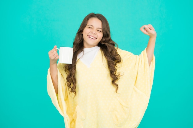 Conceito de bem-estar. Menina com cabelo comprido. Me sentindo confortável. Criança adorável em fundo turquesa. Roupa confortável para descanso de fim de semana em casa. Estilo confortável. Criança usando poncho macio, bebendo chá.