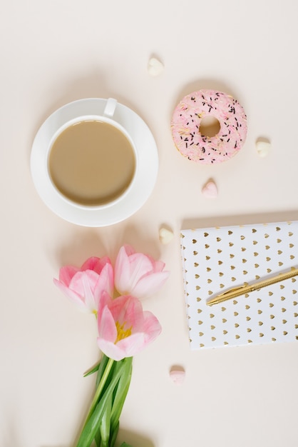 Conceito de blog de primavera. Xícara de café, donut e buquê de tulipas cor de rosa em fundo bege. Postura plana, mesa feminina com vista superior.