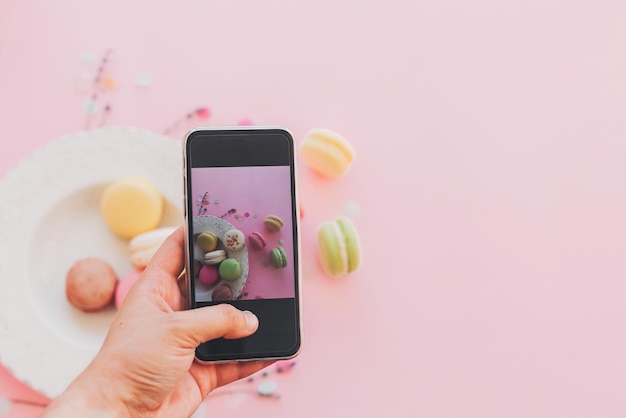 Conceito de blogging do instagram fotografia de comida plana mão segurando o telefone e tirando foto de biscoitos coloridos elegantes em prato vintage no espaço de papel rosa da moda para texto