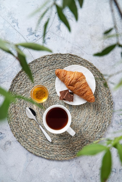 Conceito de café da manhã com uma xícara de chá mel e um croissant em uma mesa de mármore e galhos verdes closeup vista superior orientação vertical