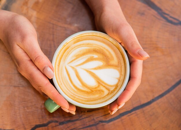 Conceito de cafeteria e café. Cappuccino de mãos femininas. Desenhe na superfície do café com leite. Desafio para artista de latte. Xícara de café de vista superior na mesa de madeira. Belo padrão de coração criado por microfoam.