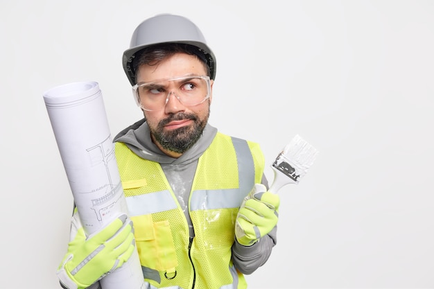 Conceito de carreira de trabalho de ocupação. o homem sério e trabalhador tem uma expressão pensativa e segura a planta para a construção do futuro pincel de pintura usa óculos de capacete de segurança e espaço em branco uniforme à direita