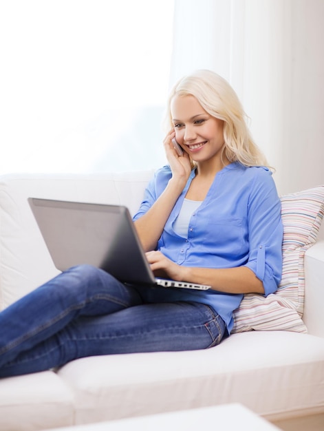 conceito de casa, tecnologia e internet - mulher sorridente com smartphone e laptop sentado no sofá em casa