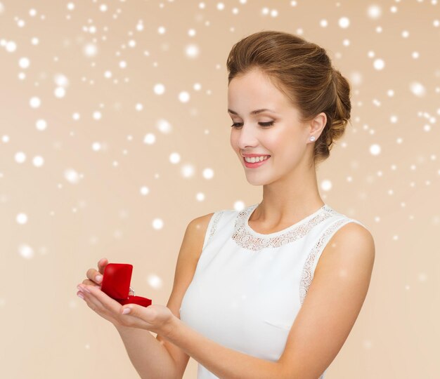 conceito de casamento, amor, noivado e felicidade - mulher sorridente no vestido branco segurando a caixa de presente vermelha com anel de diamante sobre fundo bege e neve