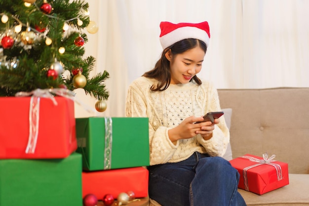 Conceito de celebração de natal jovem mulher asiática sentada na sala de estar e conversando no smartphone