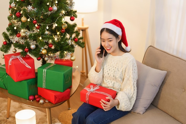 Conceito de celebração de Natal Jovem mulher asiática sentada na sala de estar e falando no smartphone