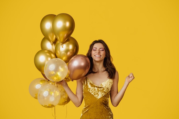 Conceito de celebração do feriado Mulher feliz em vestido segurando um monte de balões e fazendo desejo posando em fundo amarelo