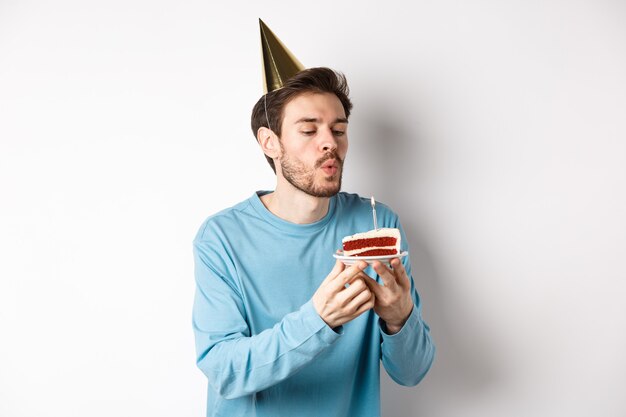 Conceito de celebração e feriados. cara bobo feliz soprando vela no bolo e fazendo desejo, em pé no chapéu de festa sobre fundo branco.