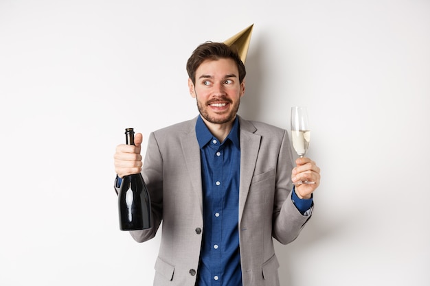 Conceito de celebração e feriados. feliz aniversário cara de terno e chapéu de festa bebendo champanhe, segurando a garrafa e o copo, sorrindo e olhando para o lado.