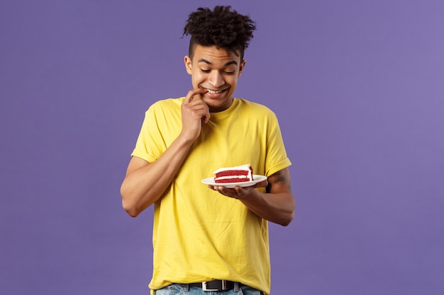 Conceito de celebração, festa e feriados. Retrato do namorado não pode resistir à tentação de comer o último pedaço de bolo, mordendo os lábios e sorrindo ansioso para comer a sobremesa, hesitando, parede roxa