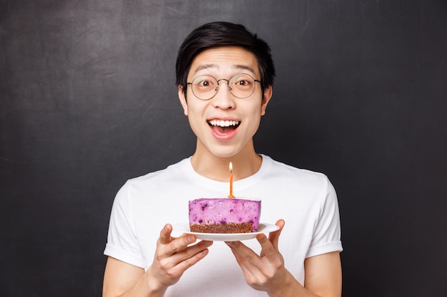 Conceito de comemoração, feriado e aniversário. retrato do close-up de espantado e feliz jovem asiático segurando bolo b-dia e sorrindo divertido, jogar festa, apague a vela para fazer desejo