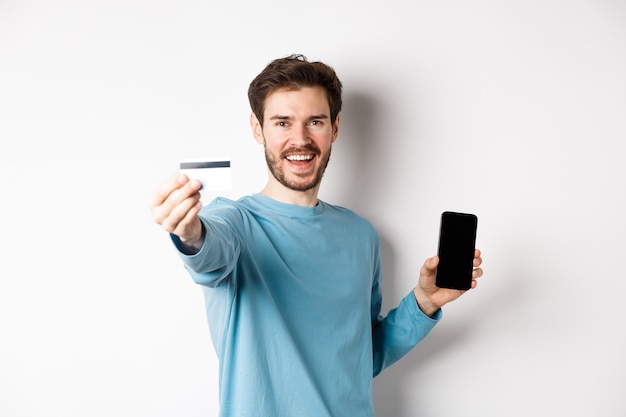 Conceito de comércio eletrônico e compras. Homem feliz, mostrando o cartão de crédito de plástico e a tela do smartphone, recomendando o aplicativo bancário, de pé sobre um fundo branco.