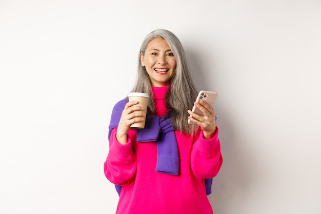 Conceito de comércio eletrônico. Linda mulher asiática sênior tomando café da xícara para viagem e usando o telefone celular, sorrindo para a câmera, fundo branco