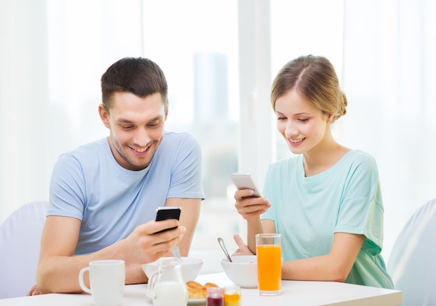 conceito de comida, casa, casal e tecnologia - casal sorridente com smartphones lendo notícias e tomando café da manhã em casa