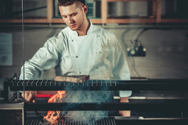 Conceito de comida jovem chef bonito em uniforme branco monitora o grau de assado e transforma a carne