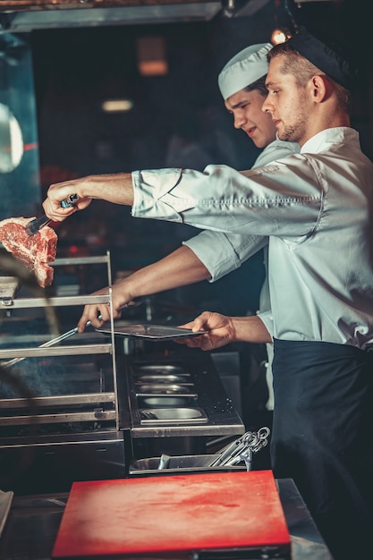 Conceito de comida. jovens chefs bonitos em uniforme branco acendem carvões e colocam carne crua marinada na grelha no interior da cozinha do restaurante. preparando o tradicional bife no forno de churrasco.