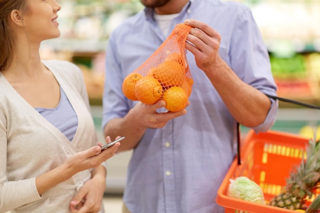 Foto conceito de compras, comida, venda, consumismo e pessoas - casal feliz com smartphone comprando laranjas em mercearia ou supermercado