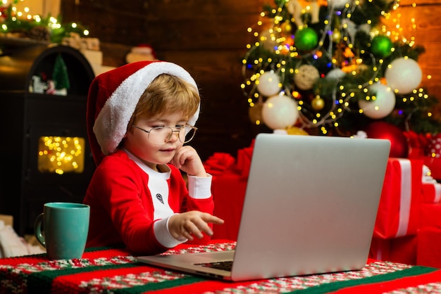 Conceito de compras de Natal. Serviço de presentes. Ajudante de Papai Noel. Criança inteligente a navegar na Internet. Traje e chapéu de Papai Noel de menino pequeno. Menino com laptop perto da árvore de Natal. Compre presentes de natal online