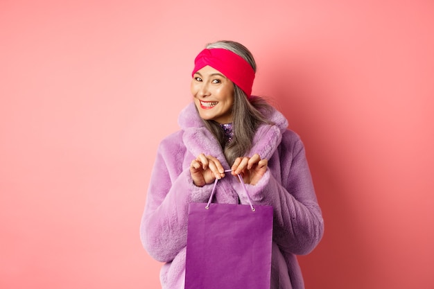 Conceito de compras e moda. a avó elegante comprou um presente e sorriu, segurando uma bolsa roxa nas mãos, em pé sobre um fundo rosa