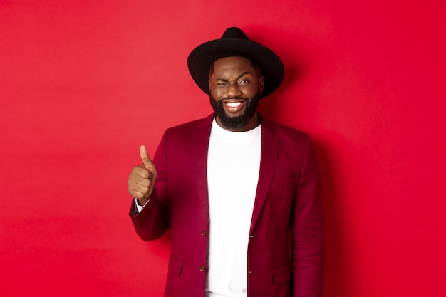 Conceito de compras e pessoas de Natal. Homem negro barbudo bonito com blazer de festa mostrando o polegar para cima, piscando e sorrindo para a câmera, recomendando promoção, fundo vermelho