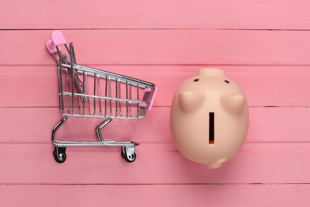 Conceito de compras, economia. cofrinho com carrinho de supermercado na superfície de madeira rosa. vista do topo