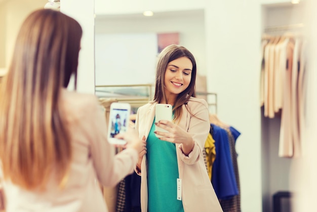 Foto conceito de compras, moda, estilo, tecnologia e pessoas - mulher feliz com smartphone tirando selfie de espelho na loja de roupas