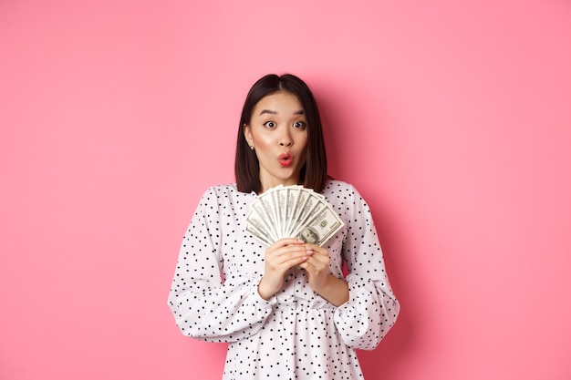 Conceito de compras. Mulher asiática animada segurando dinheiro, ofegando e espantada e olhando para a câmera, em pé sobre rosa.