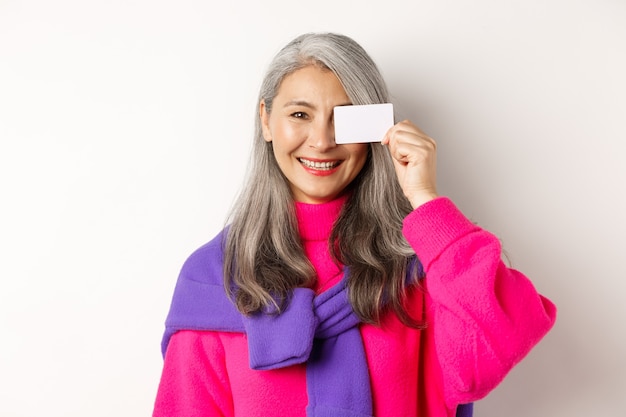 Conceito de compras. Mulher asiática sênior elegante sorrindo e mostrando o cartão de crédito de plástico, pagando sem contato, em pé sobre um fundo branco.