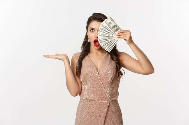 Conceito de compras. Mulher chocada segurando dinheiro perto do rosto, usando um vestido elegante, mostrando dólares com expressão de espanto, em pé sobre um fundo branco.