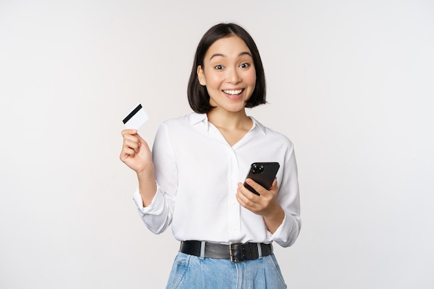Conceito de compras on-line Imagem de jovem asiática moderna segurando cartão de crédito e compra de smartphone com aplicativo de smartphone pagando sem contato em pé sobre fundo branco