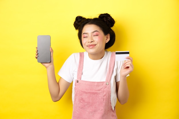 Conceito de compras online. Feliz menina asiática mostrando a tela do smartphone e o cartão de crédito vazio, pagando sem contato, parado no amarelo