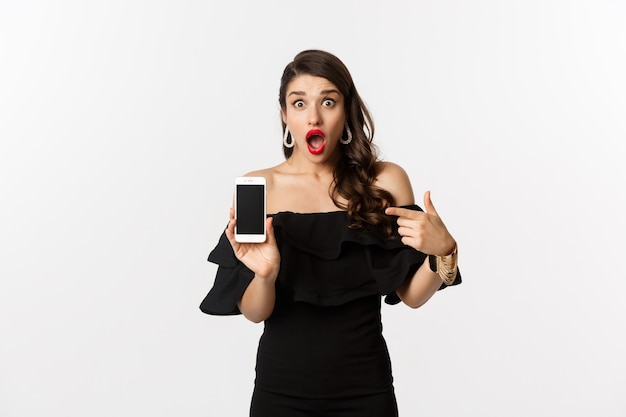 Conceito de compras online. Mulher elegante em vestido preto apontando o dedo para a tela do smartphone, mostrando o aplicativo, em pé sobre um fundo branco.