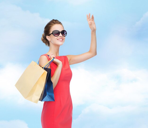 conceito de compras, venda, natal e férias - mulher elegante sorridente em vestido vermelho e óculos de sol com sacolas de compras acenando com a mão