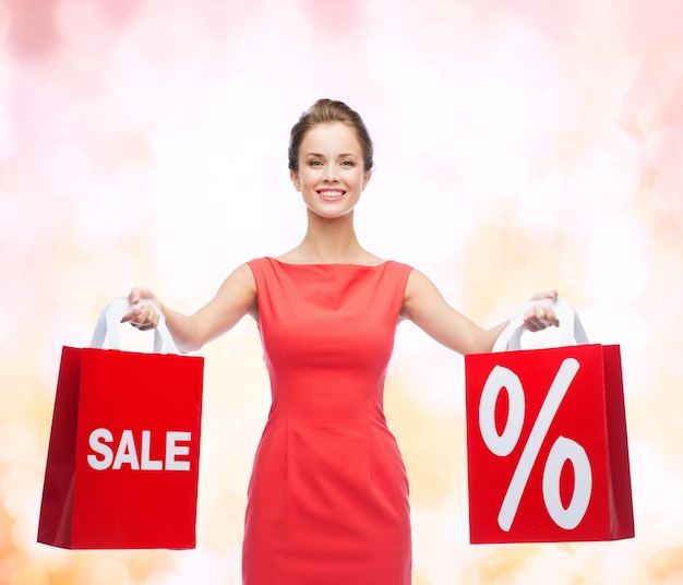 conceito de compras, venda, presentes, natal e férias - jovem sorridente de vestido vermelho com sacolas de compras com porcentagem e sinal de venda