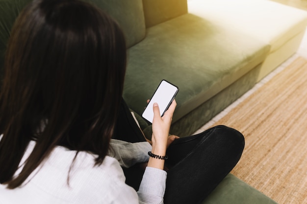 Conceito de comunicação com a garota negra ouve a menina sentada no sofá verde e segurando o smartphone na mão com uma tela branca vazia, raio de sol na frente