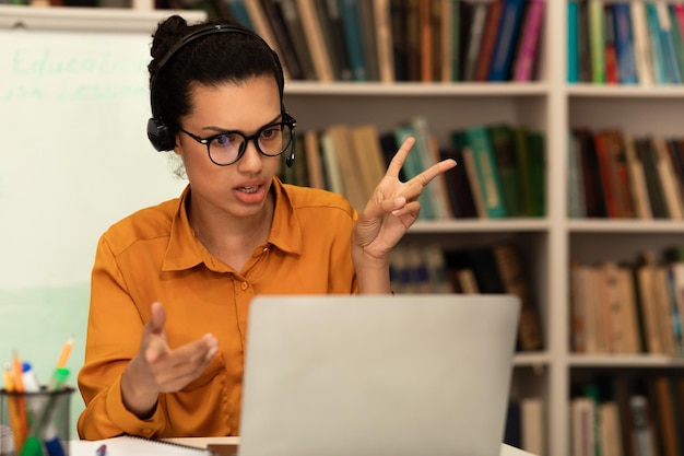 Conceito de comunicação distante remota professora de raça mista em fones de ouvido falando e gesticulando na webcam do laptop