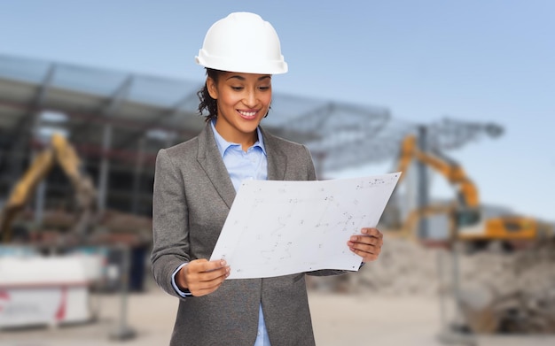 conceito de construção, desenvolvimento, construção e arquitetura - empresária afro-americana sorridente no capacete branco olhando para a planta