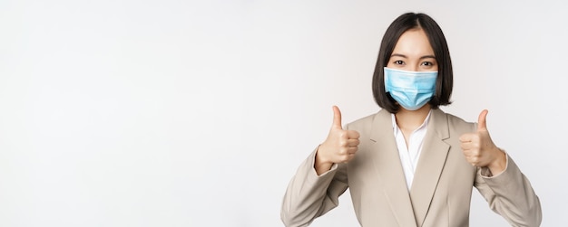 Conceito de coronavírus e local de trabalho imagem do trabalhador da empresa vendedora asiática em máscara médica mostrando os polegares para cima sinal sorrindo fundo branco satisfeito