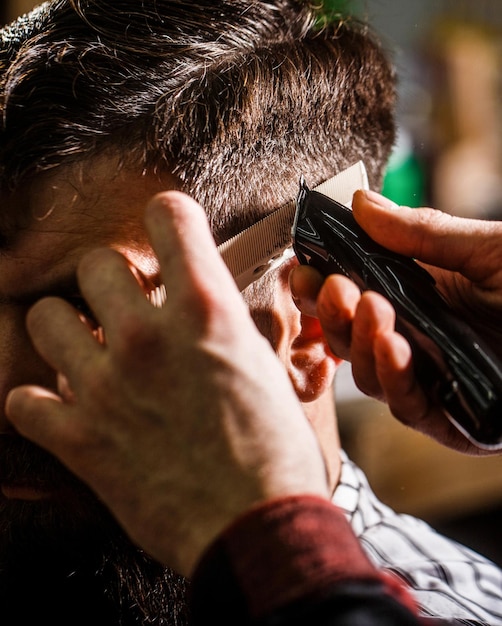 Conceito de corte de cabelo homem visitando cabeleireiro na barbearia mãos de barbeiro com máquina de cortar cabelo close-up homem barbudo na barbearia