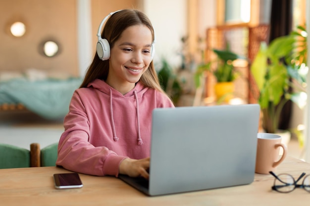 Conceito de crianças e tecnologia Adolescente feliz sentada à mesa e usando laptop usando fones de ouvido sem fio copiam espaço