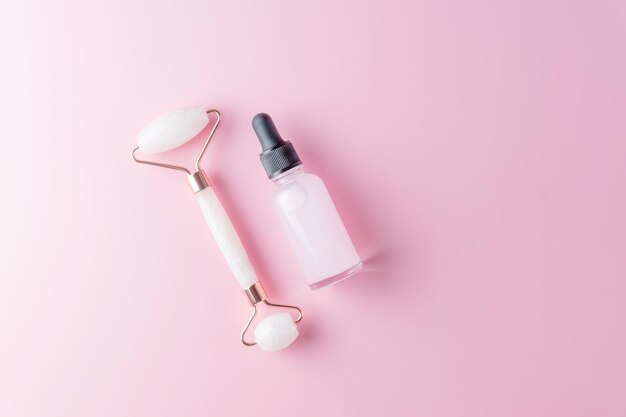 Conceito de cuidados com a pele. Frasco de produto comético, soro ou óleo. Rolo de quartzo. Cosméticos e ferramentas em fundo rosa