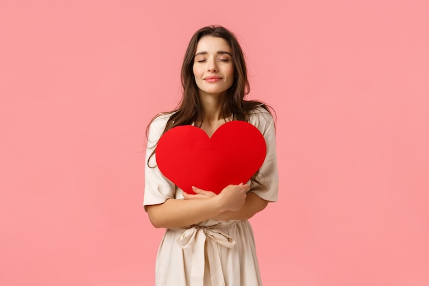 Conceito de cuidados, ternura e beleza. Concurso e adorável jovem namorada em vestido de pé sobre a parede rosa, abraçando o cartão grande coração com olhos fechados e sorriso fofo, parede rosa em pé